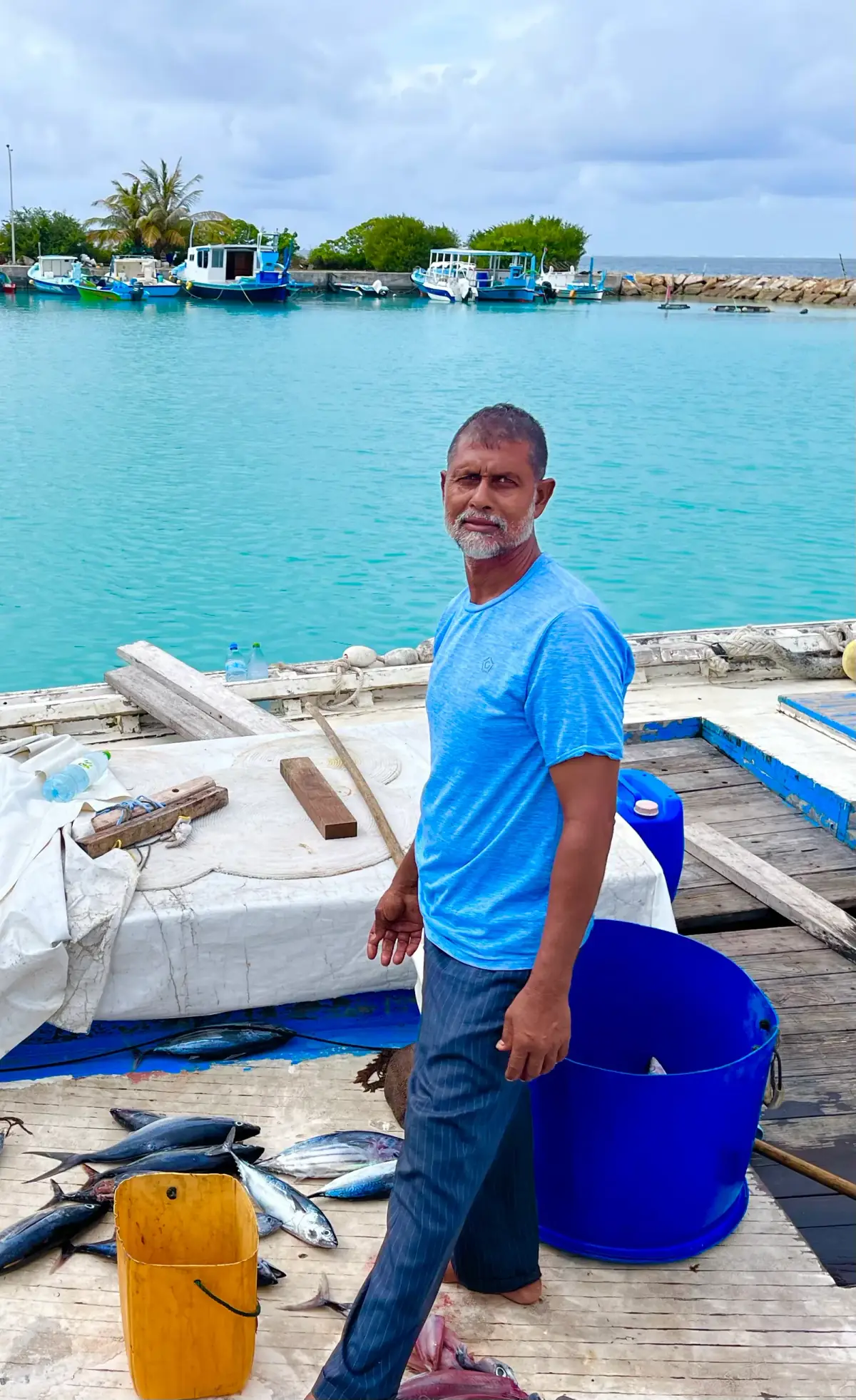 Fischer in blauem Shirt auf Holzsteg mit frischem Fang, Fischerboote im türkisfarbenen Wasser, tropische Küstenszene