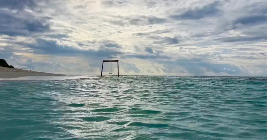 Türkisblaues Meer mit verlassenem Schaukelgerüst im Wasser, bewölkter Himmel und Inselküste im Hintergrund