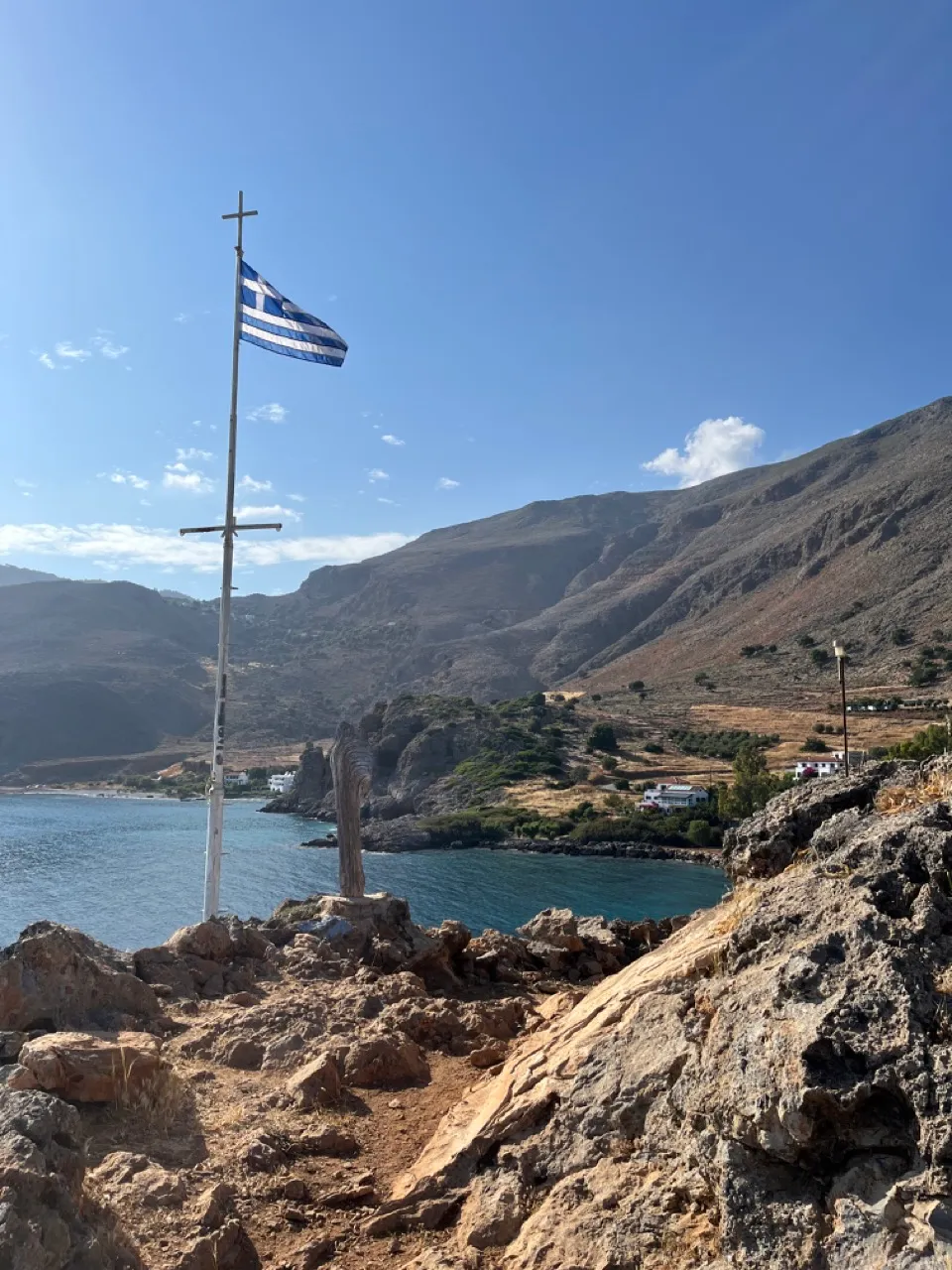 Weißes Holzkreuz mit griechischer Flagge auf felsiger Küstenklippe über türkisblauem Meer