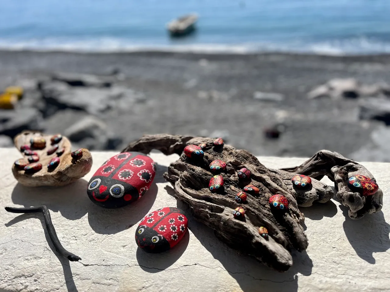 Bunte Kunstmarienläfer aus Holz und bemalte Steine liegen am Strand neben Treibholz, im Hintergrund das Meer mit einem Boot