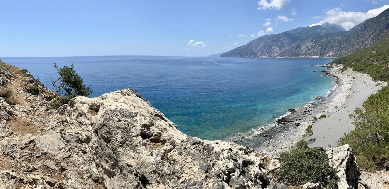 Blick von Felsen auf türkisklare Bucht mit Kiesstrand, umgeben von bewaldeten Bergen unter blauem Himmel