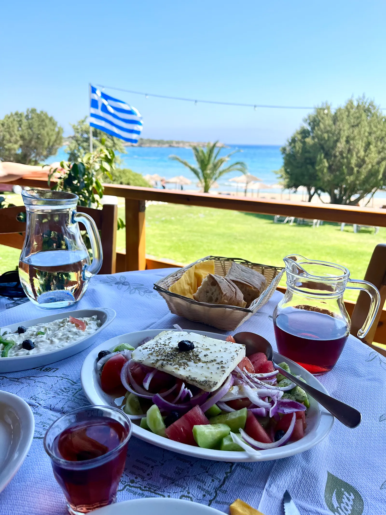 Festliche Mahlzeit mit Feta-Salat und Rotwein auf Terrasse mit Meerblick, griechische Flagge im Hintergrund
